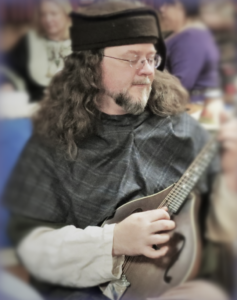 A man with long, brown hair, wearing a medieval hood and hat, eyes closed and playing a mandolin.