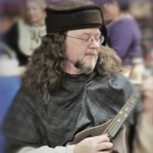 A man with long, brown hair, wearing a medieval hood and hat, eyes closed and playing a mandolin.